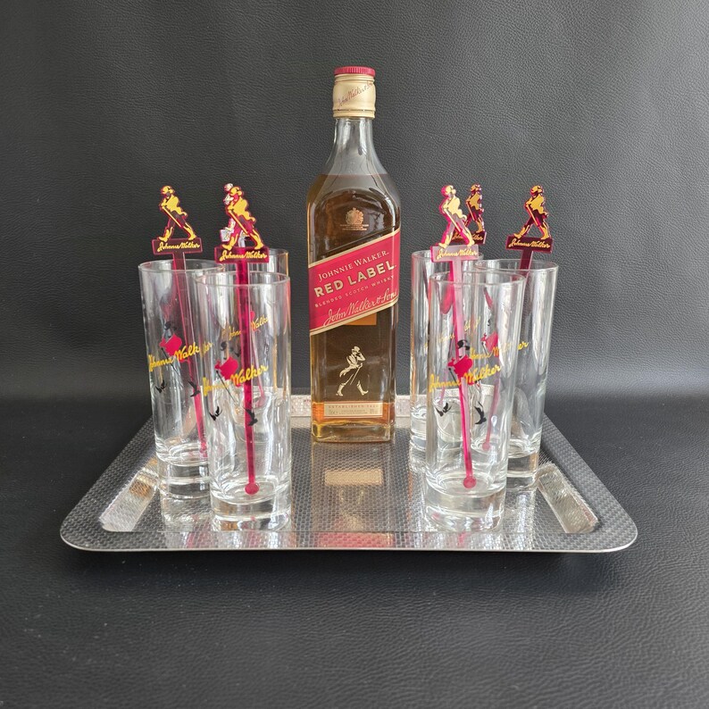 May include: A silver serving tray holds a bottle of Johnnie Walker Red Label whisky, six tall glasses, and cocktail stirrers. The glasses and stirrers feature a red and gold logo. The bottle is amber-coloured.