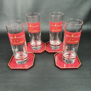 May include: Four clear glass tumblers with the words "Keep Walking Johnnie Walker" printed on them. Each glass is sitting on a red coaster with a gold border.