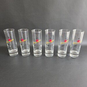 May include: Six clear shot glasses with a red heart and yellow text reading "Johnnie Walker". The cylindrical glasses are arranged on a dark surface.