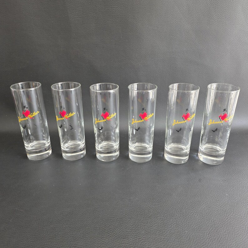 May include: Six clear shot glasses with a red heart and yellow text reading "Johnnie Walker". The cylindrical glasses are arranged on a dark surface.