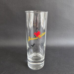 May include: A clear, tall glass featuring the "Johnnie Walker" logo in yellow script, a red figure, and black accents. The cylindrical glass has a slightly flared top, ideal for serving drinks.