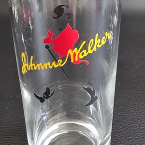 May include: A clear glass featuring the Johnnie Walker logo. The logo shows a red silhouette of a man with a black hat and cane, with the words "Johnnie Walker" in yellow cursive. Two black butterfly shapes are at the bottom.