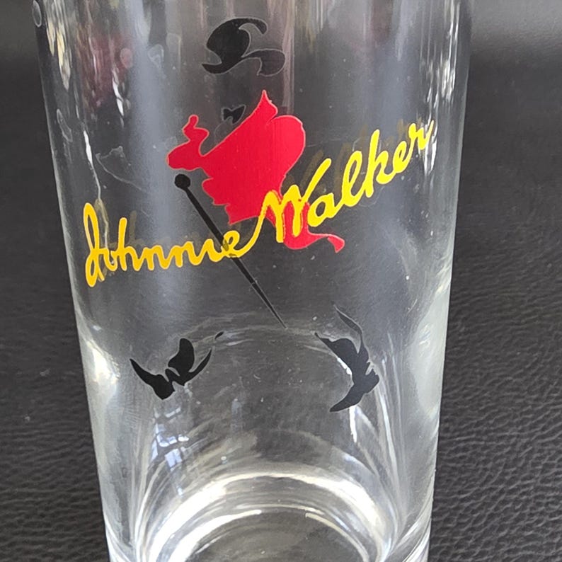 May include: A clear glass featuring the Johnnie Walker logo. The logo shows a red silhouette of a man with a black hat and cane, with the words "Johnnie Walker" in yellow cursive. Two black butterfly shapes are at the bottom.