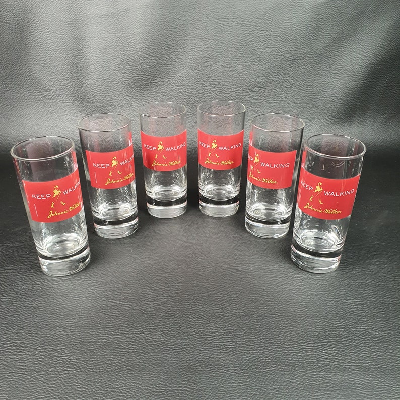 May include: Six clear shot glasses with a red band featuring the words "KEEP WALKING" and the "Johnnie Walker" logo. The glasses are cylindrical and sit on a dark surface.