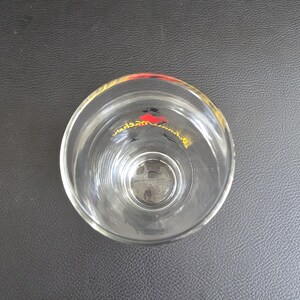 May include: A clear glass tumbler with a flared rim. The glass features a design in red, black, and yellow near the top. The glass is empty and rests on a dark grey surface.