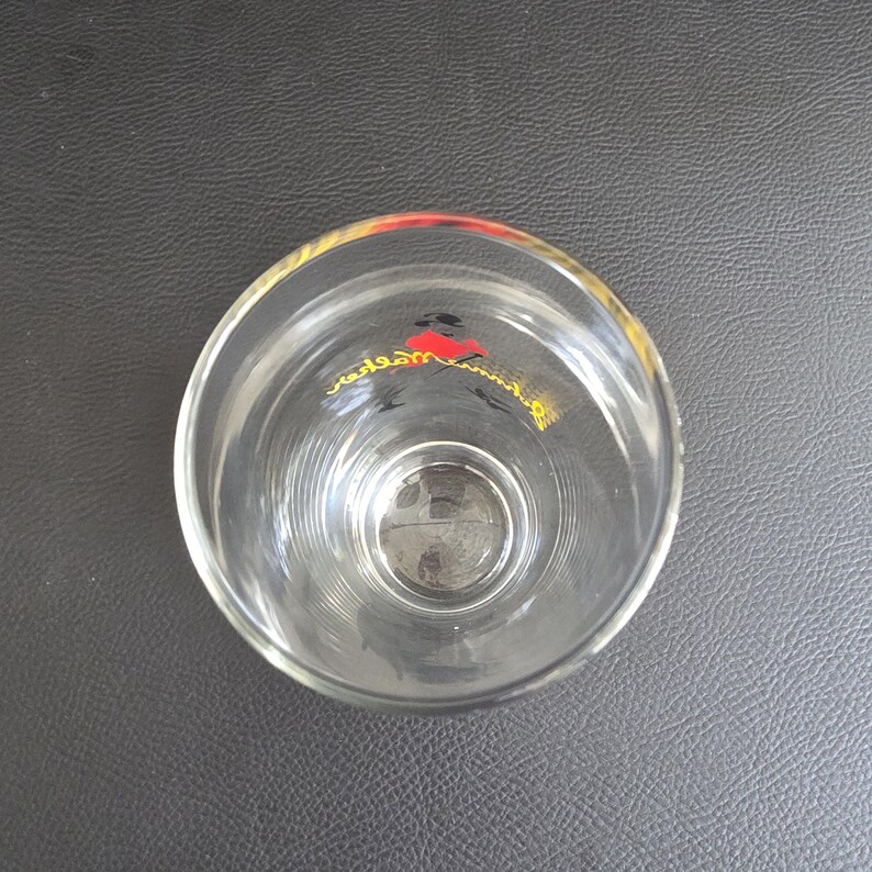 May include: A clear glass tumbler with a flared rim. The glass features a design in red, black, and yellow near the top. The glass is empty and rests on a dark grey surface.