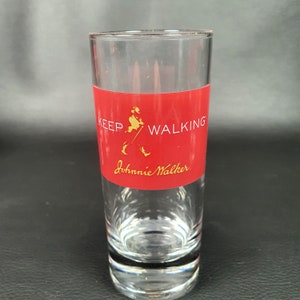 May include: A clear shot glass with a red band featuring the text "KEEP WALKING" and the "Johnnie Walker" logo in gold. The glass is cylindrical and has a thick base.