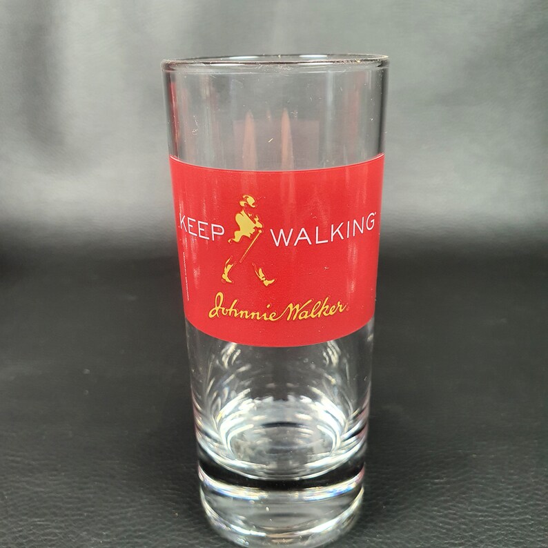 May include: A clear shot glass with a red band featuring the text "KEEP WALKING" and the "Johnnie Walker" logo in gold. The glass is cylindrical and has a thick base.