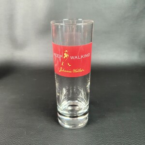 May include: A clear glass shot glass with a red band around the middle. The band has the Johnnie Walker logo and the text "Keep Walking".