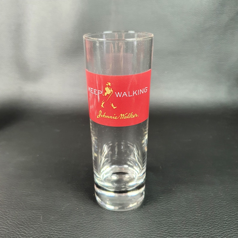 May include: A clear glass shot glass with a red band around the middle. The band has the Johnnie Walker logo and the text "Keep Walking".