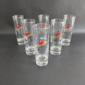 May include: Six clear, cylindrical drinking glasses. Each glass has a red heart and a yellow logo with the text "Johnnie Walker". The glasses are arranged on a dark surface.