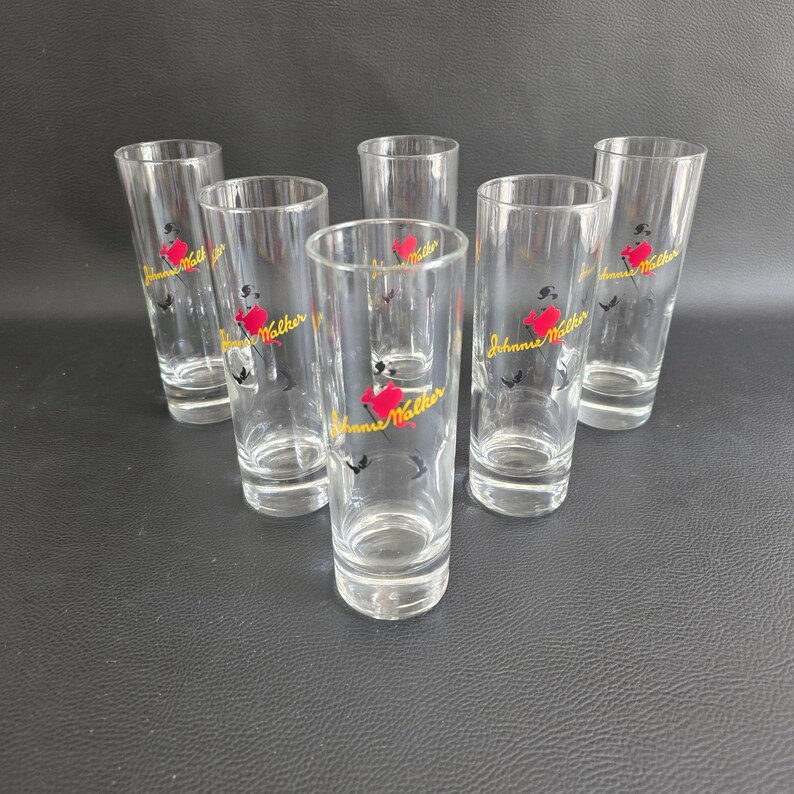 May include: Six clear, cylindrical drinking glasses. Each glass has a red heart and a yellow logo with the text "Johnnie Walker". The glasses are arranged on a dark surface.