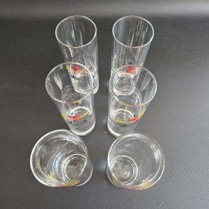 May include: Six clear glass shot glasses with gold rims and a red and yellow logo. The cylindrical glasses are empty. The shot glasses are arranged on a dark surface. Each glass is approximately 10 cm tall.