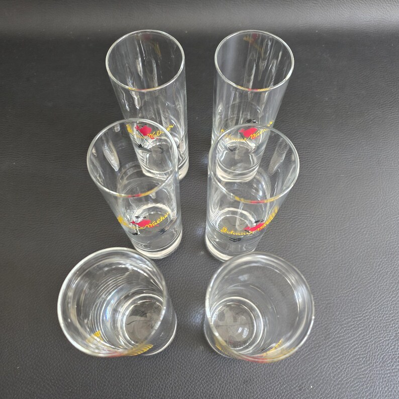May include: Six clear glass shot glasses with gold rims and a red and yellow logo. The cylindrical glasses are empty. The shot glasses are arranged on a dark surface. Each glass is approximately 10 cm tall.