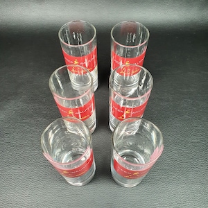 May include: Six clear shot glasses with a red band featuring gold text that reads "Keep Walking Johnnie Walker". The shot glasses are arranged in a hexagonal pattern on a dark surface.
