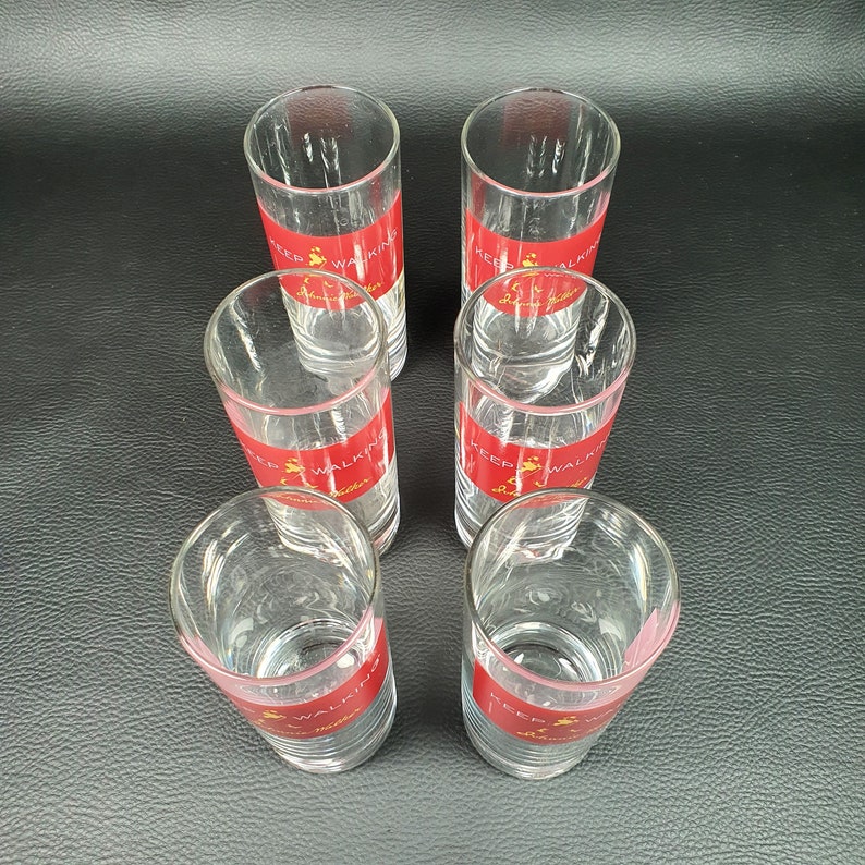 May include: Six clear shot glasses with a red band featuring gold text that reads "Keep Walking Johnnie Walker". The shot glasses are arranged in a hexagonal pattern on a dark surface.