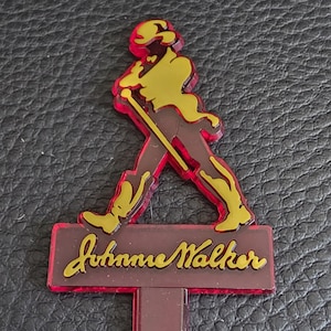 May include: A Johnnie Walker bar accessory in red, yellow, and brown. It features the Striding Man figure in yellow, outlined in red, above a brown rectangular base with the Johnnie Walker logo in yellow script.