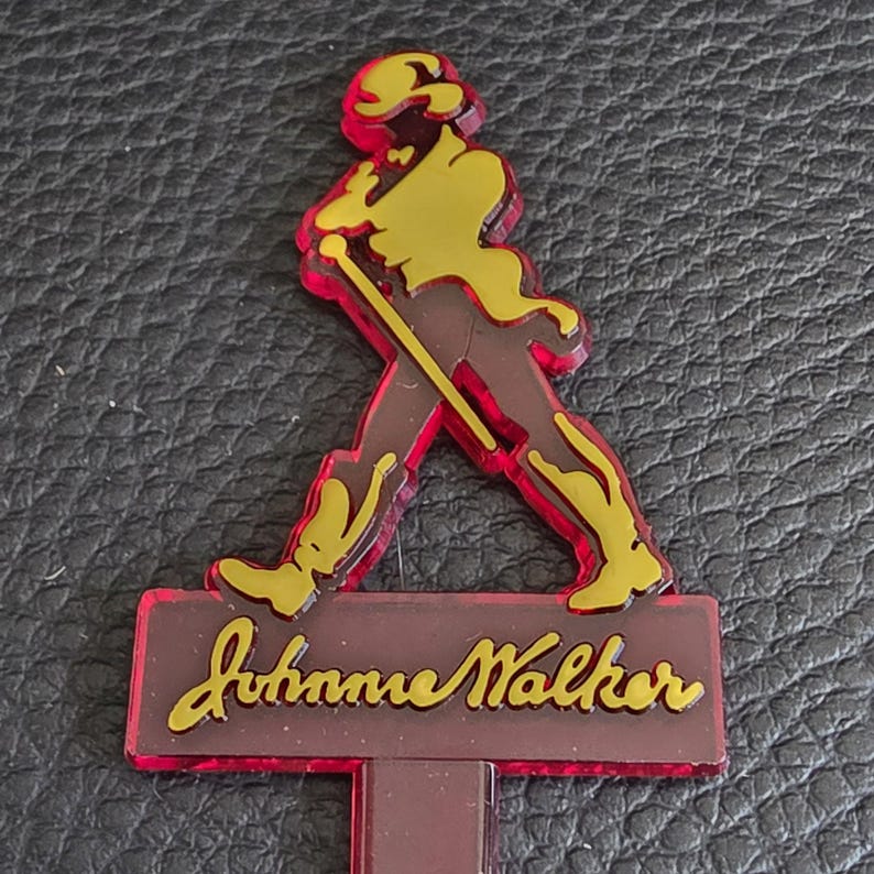 May include: A Johnnie Walker bar accessory in red, yellow, and brown. It features the Striding Man figure in yellow, outlined in red, above a brown rectangular base with the Johnnie Walker logo in yellow script.