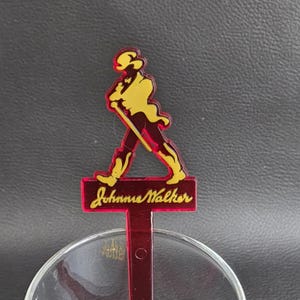 May include: A red and yellow cocktail stirrer with the Johnnie Walker logo. The logo shows a man in a top hat walking with a cane. The Johnnie Walker text is in a cursive font. The stirrer is positioned over a clear glass.
