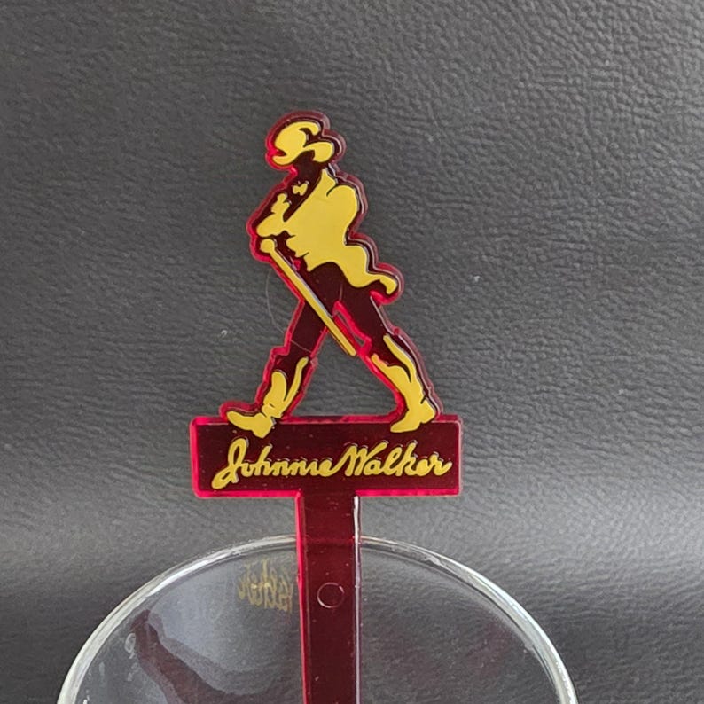 May include: A red and yellow cocktail stirrer with the Johnnie Walker logo. The logo shows a man in a top hat walking with a cane. The Johnnie Walker text is in a cursive font. The stirrer is positioned over a clear glass.