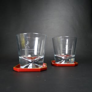May include: Two clear glass whiskey tumblers with the Johnnie Walker logo etched on the side, each sitting on a red coaster with a gold rim.