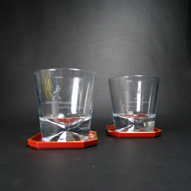 May include: Two clear glass whiskey tumblers with the Johnnie Walker logo etched on the side, each sitting on a red coaster with a gold rim.