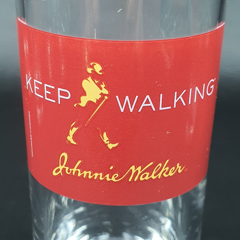 May include: A clear glass with a red band featuring the text "KEEP WALKING" and the "Johnnie Walker" logo in gold. The logo depicts a man walking with a cane. The glass is designed for beverages.