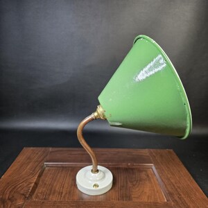 May include: A wall-mounted lamp featuring a conical green metal shade attached to a curved copper arm. The base is a circular white ceramic fixture with a single central screw, designed for mounting to a flat surface with a metallic finish.