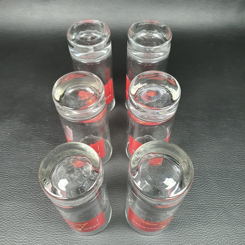 May include: Six clear, cylindrical drinking glasses with a red band around the middle. The glasses are arranged in a circular pattern, with the bottoms facing the viewer. The red band has white text. The glasses are on a black surface.