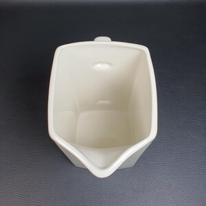 May include: A cream-colored, square-shaped creamer with a spout and a small handle. The creamer has a minimalist design and is made of ceramic. The interior is smooth, and the exterior has a slightly textured surface.