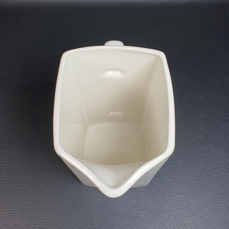 May include: A cream-colored, square-shaped creamer with a spout and a small handle. The creamer has a minimalist design and is made of ceramic. The interior is smooth, and the exterior has a slightly textured surface.