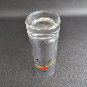 May include: A clear, cylindrical glass with a slightly flared top. The glass has a logo in red and yellow near the base. The glass is empty and sits on a dark surface.