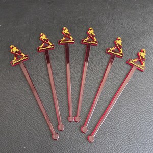 May include: Six red plastic cocktail stirrers. Each stirrer features a rectangular top with the text "Johnnie Walker" and a gold figure of a man walking. The stirrers are arranged on a dark surface.