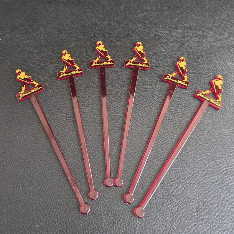 May include: Six red plastic cocktail stirrers. Each stirrer features a rectangular top with the text "Johnnie Walker" and a gold figure of a man walking. The stirrers are arranged on a dark surface.
