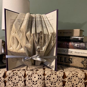 May include: A book with pages folded into a 3D design of a heart. The book is on a table with a crocheted tablecloth. There are other books stacked behind the book with the heart design. The book with the heart design has a purple spine and a silver ribbon tied around it.