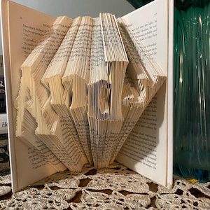 May include: A handmade book sculpture with the word "HOME" folded into the pages. The open book displays the word in a 3D design. The pages are aged, and the book is set on a decorative surface.