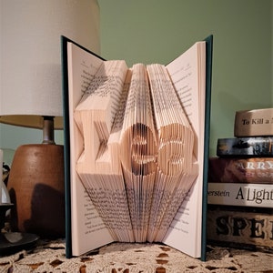 Puede incluir: Un libro abierto con las páginas plegadas para formar la palabra "LOVE". Las letras esculpidas se destacan sobre el fondo de las páginas. El libro está sobre una mesa con un tapete de encaje.