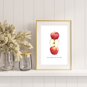 May include: A framed print with a gold border features a watercolor illustration of three red apples, one whole and one sliced open. The text "THE APPLE OF MY EYE." is below the apples. The print is displayed on a white shelf.