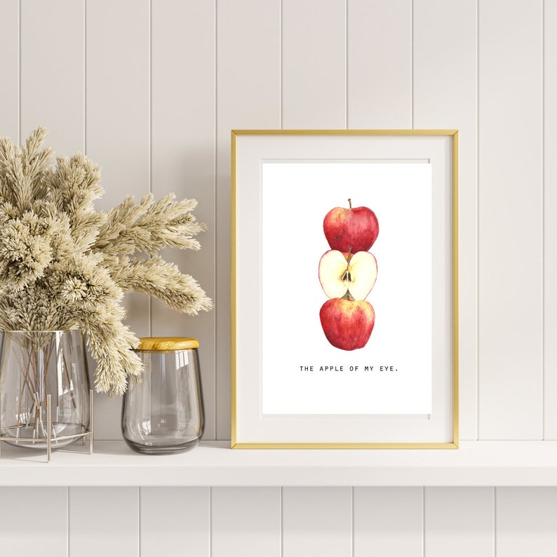 May include: A framed print with a gold border features a watercolor illustration of three red apples, one whole and one sliced open. The text "THE APPLE OF MY EYE." is below the apples. The print is displayed on a white shelf.