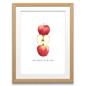 May include: A framed print featuring a watercolor illustration of three red apples stacked vertically. The middle apple is sliced in half, revealing the interior. The text "THE APPLE OF MY EYE." is below the apples.