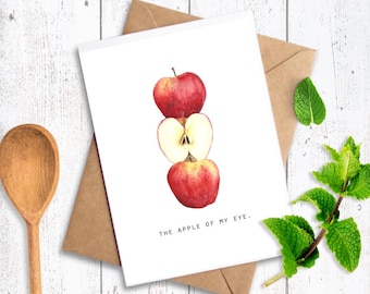 May include: A greeting card with a watercolor illustration of three red apples, one halved to show the inside. The card is on a brown envelope, with the text "THE APPLE OF MY EYE." A wooden spoon and sprigs of mint are also visible.