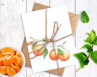 May include: A greeting card featuring a watercolour illustration of oranges tied with twine, placed on a brown envelope. A small white bowl of orange segments and sprigs of mint are on a white wooden surface.