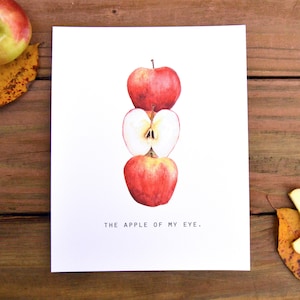 May include: A print featuring a watercolor illustration of three stacked red apples, one sliced open to reveal the core. The text "THE APPLE OF MY EYE." is below the apples. The print is on a wooden surface with an apple and leaves.