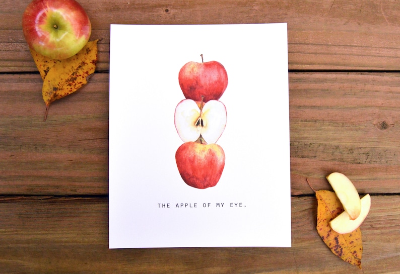May include: A print featuring a watercolor illustration of three stacked red apples, one sliced open to reveal the core. The text "THE APPLE OF MY EYE." is below the apples. The print is on a wooden surface with an apple and leaves.