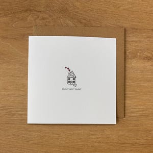 May include: A white greeting card with a hand-drawn house illustration and the phrase "Home sweet home!" in black ink. The card is paired with a brown envelope, set against a wooden background.
