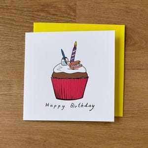 Op de afbeelding: Een witte wenskaart met een cupcake-illustratie, een brandende kaars en de tekst "Happy Birthday". De cupcake heeft een rode liner, bruine cake, witte glazuur en een pen en inktpot. Achter de kaart zit een gele envelop.