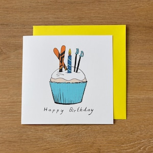 Può includere: Un biglietto di auguri bianco con un'illustrazione di cupcake blu, sci arancioni e candele. La carta ha le parole "Happy Birthday" scritte in nero. Una busta gialla è dietro la carta.