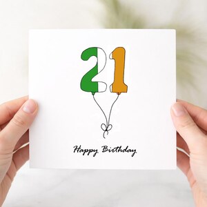 May include: A white square greeting card featuring a hand-drawn illustration of the number 21 shaped like balloons. The number 2 is coloured green and white, while the number 1 is orange. Black strings tie the balloons together, with the text Happy Birthday.