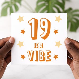 May include: A white card with the text "19 IS A VIBE" in orange, surrounded by orange and yellow stars. The card is held by hands with dark skin.