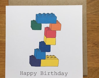 Puede incluir: Una tarjeta de felicitación cuadrada con un gráfico del número dos construido con bloques de construcción de plástico de colores azul, verde, naranja, rojo y amarillo. El texto Happy Birthday está impreso en negro con fuente de máquina de escribir.